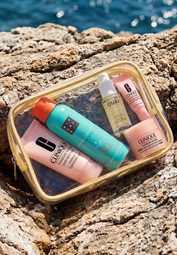 Trousse de toilette transparente avec des soins Clinique, un spray solaire bleu SPF 30 et de l'huile pour cheveux OUAI reposant sur des rochers texturés au bord de l'eau.