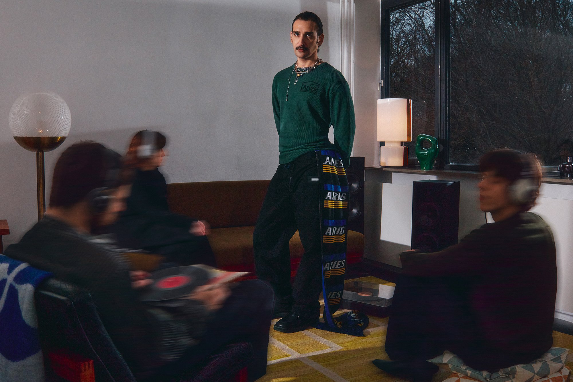 Una persona con un felpa verde e pantaloni neri sta in piedi con sicurezza, mentre altre tre ascoltano musica con le cuffie in una stanza accogliente.