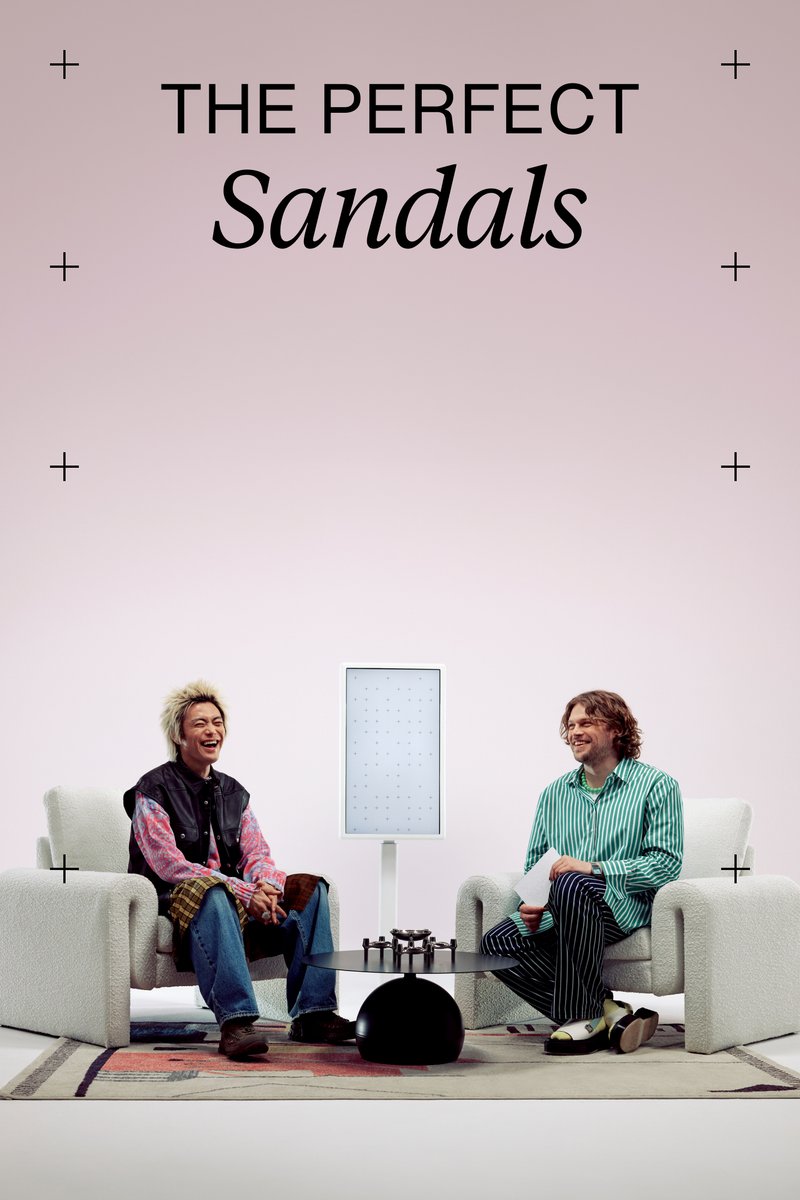 Dos personas sentadas en sillones de terciopelo, sonriendo y conversando frente a una pequeña mesa de café negra y una vitrina iluminada detrás de ellos, con un fondo rosa.
