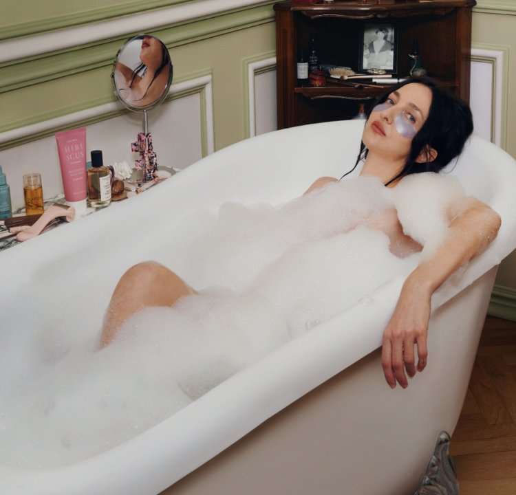 Vit fristående badkar fyllt med bubblor; en person med mörkt hår kopplar av inuti medan hen bär silvriga ögonlappar. Bakgrunden har vintageinredning.