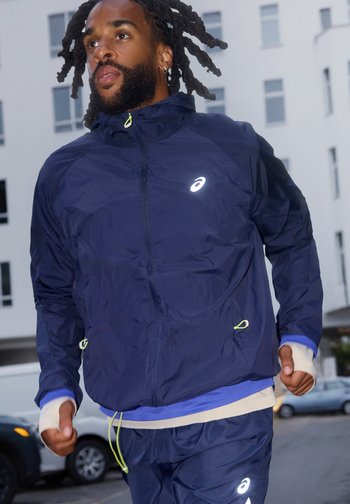 Αθλητικός άνδρας με κοτσιδάκια τρέχει έξω φορώντας μπλε φόρμα με ανακλαστικές λεπτομέρειες λογότυπου σε αστικό περιβάλλον.