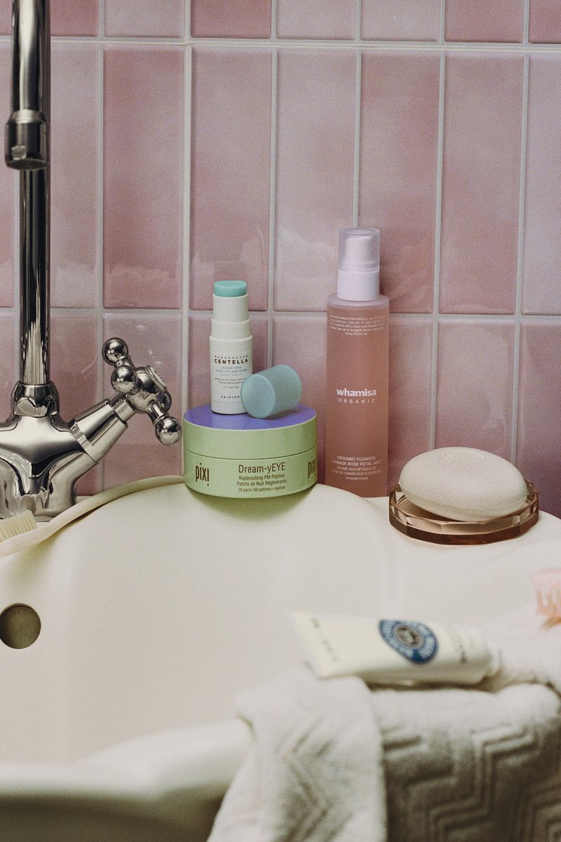 Waschbecken im Badezimmer mit Vintage-Armatur, rosa gefliester Wand, Hautpflegeprodukte einschließlich Pixi Dream-yEYE und Whamisa Spray, sowie gefaltete Handtücher.