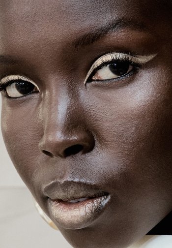 Close-up van een gezicht met een donkere huidtint, met metallic gouden oogschaduw en bijpassende metallic lippenstift, waarbij de nadruk ligt op de oogmake-up en lippen.