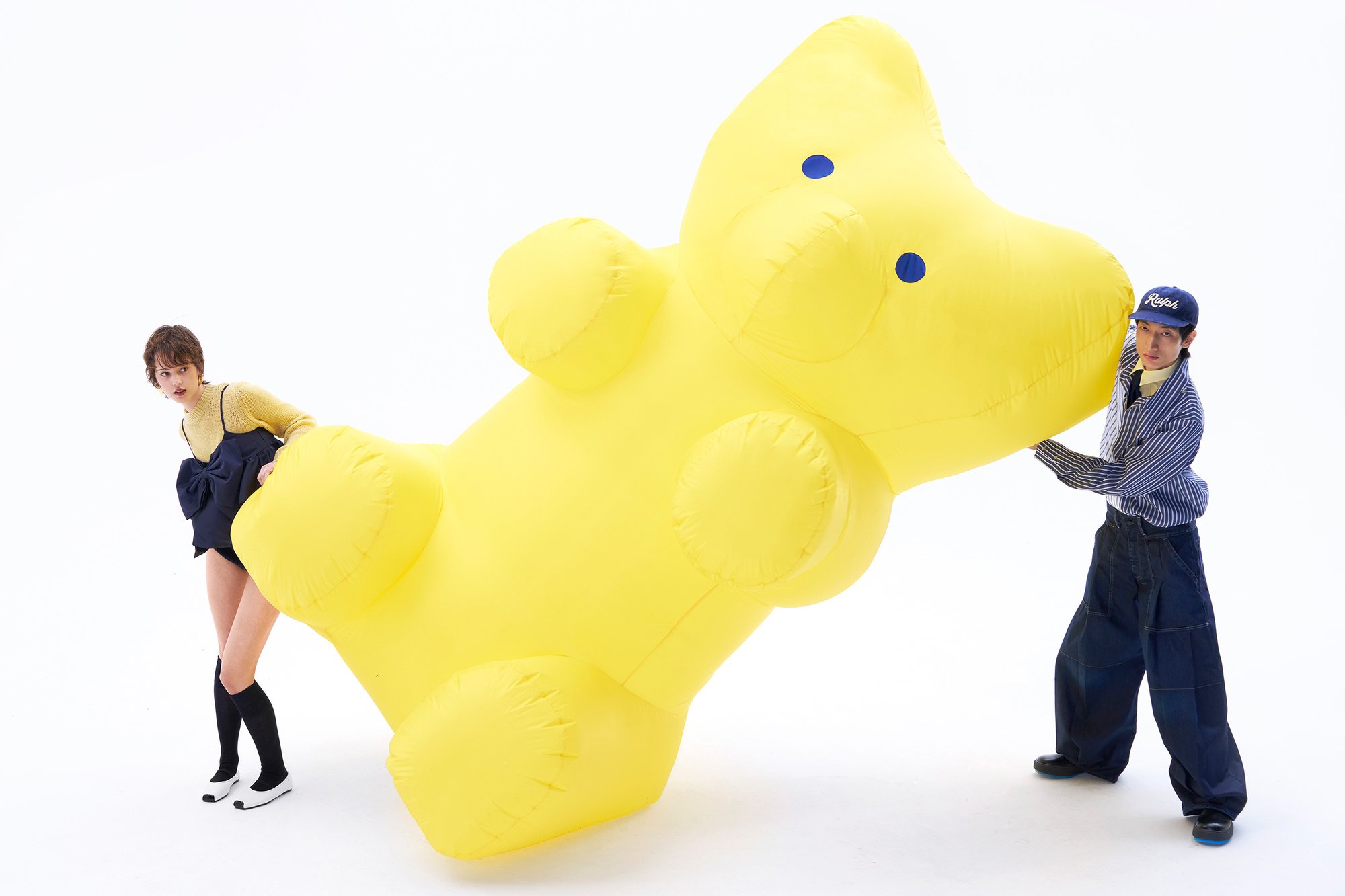 Una mujer con un suéter amarillo y un vestido azul marino sostiene un gran oso inflable amarillo, mientras un hombre con una camiseta a rayas y una gorra ayuda a cargarlo.
