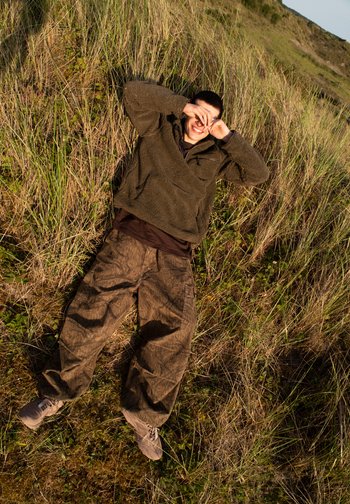 Een persoon ligt in het hoge gras, glimlachend en zijn ogen bedekkend met de handen, gekleed in een donkere jas en camouflagebroek, in een natuurlijke omgeving.