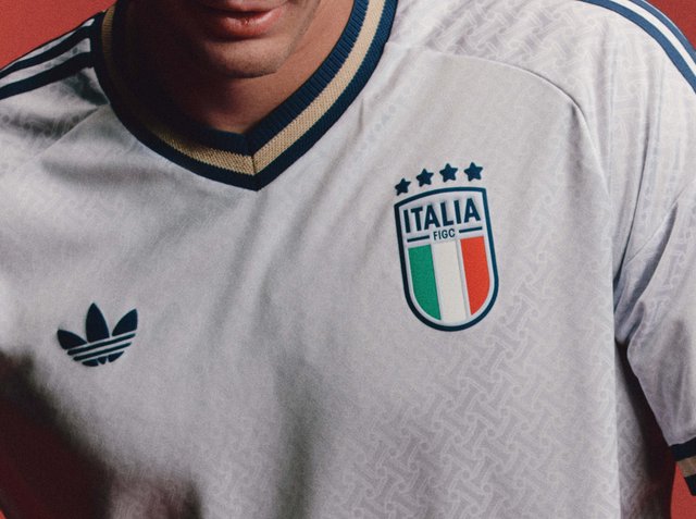 Man draagt een wit Italiaans voetbalshirt met een groen-wit-rood schildembleem en het Adidas-logo op de borst, tegen een rode achtergrond.
