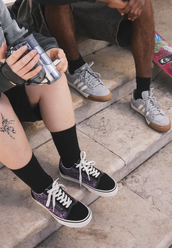Two people sitting on stone steps; one holds a camcorder, the other rests hands on knees; skateboard nearby on the floor.