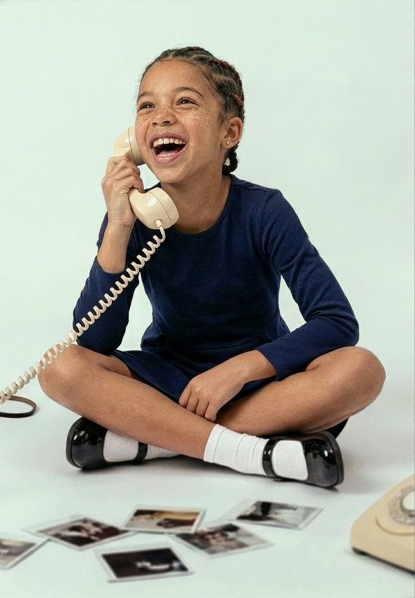Jeune fille aux cheveux tressés riant, assise en tailleur sur le sol, tenant un combiné téléphonique rétro beige près de photos éparpillées.