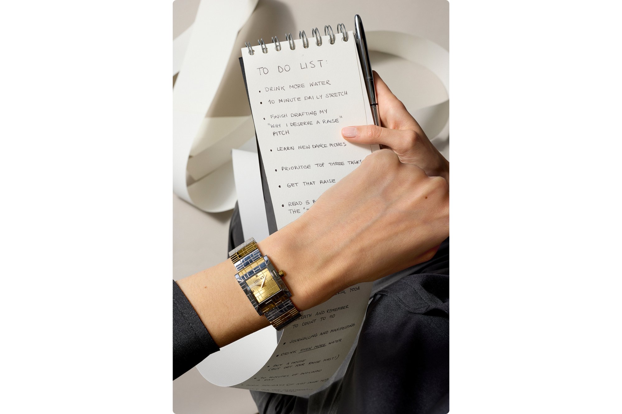 Una mano tiene un bloc note con una lista di cose da fare, mostrando compiti come "bere più acqua" e "ottenere quell’aumento," mentre indossa un orologio di moda.