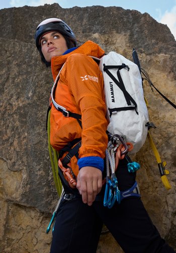 Woman climber wearing helmet, orange jacket, harness with carabiners, and white Mammut backpack stands against rocky cliff looking upwards.