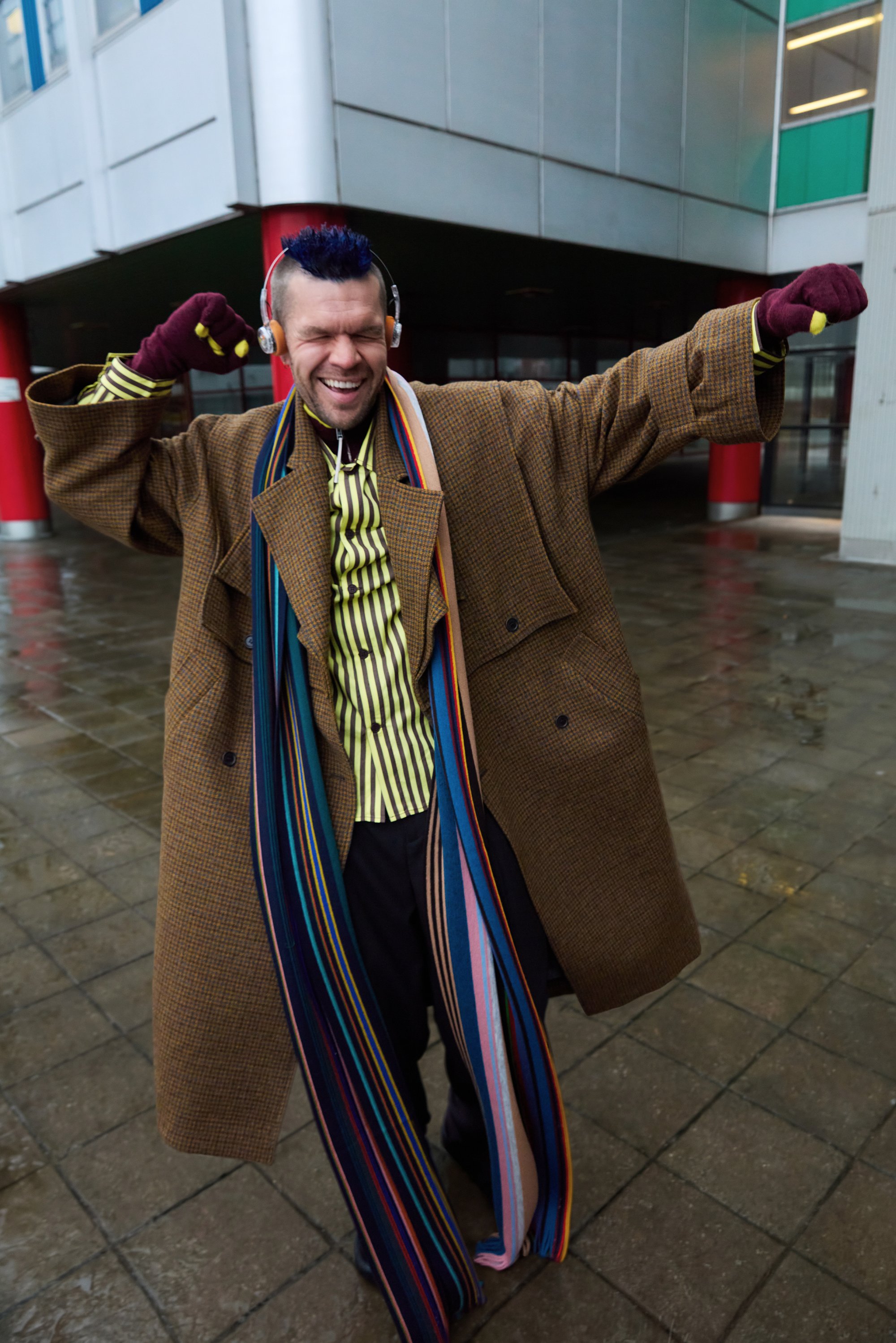 Pitkä ruskea ruudullinen ylävaate, vihreä ja keltarahkuinen raidallinen paita, värikäs raidallinen huivi ja harmaat housut. Henkilö poseeraa ulkona kuulokkeiden kanssa.