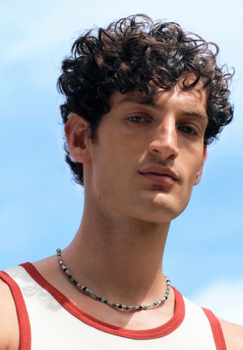 Jeune homme aux cheveux noirs bouclés portant un débardeur blanc avec des bordures rouges et un collier de perles, sur fond de ciel bleu clair.