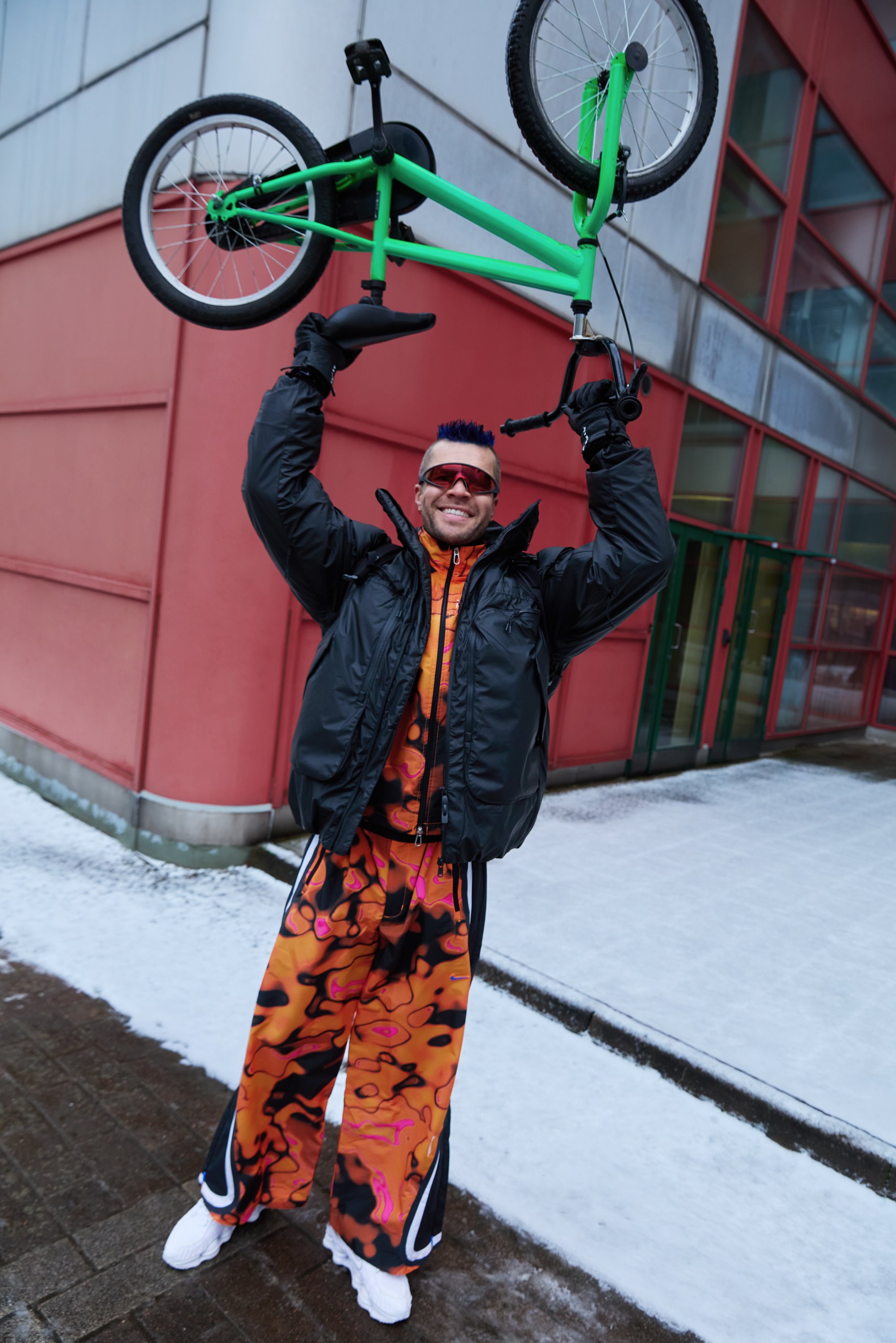 BMX-pyörä pidetty ylhäällä, jossa kirkkaanvihreä runko. Henkilöllä on musta toppatakki ja värikkäät ylipitkät housut, joissa on abstrakti kuvio.