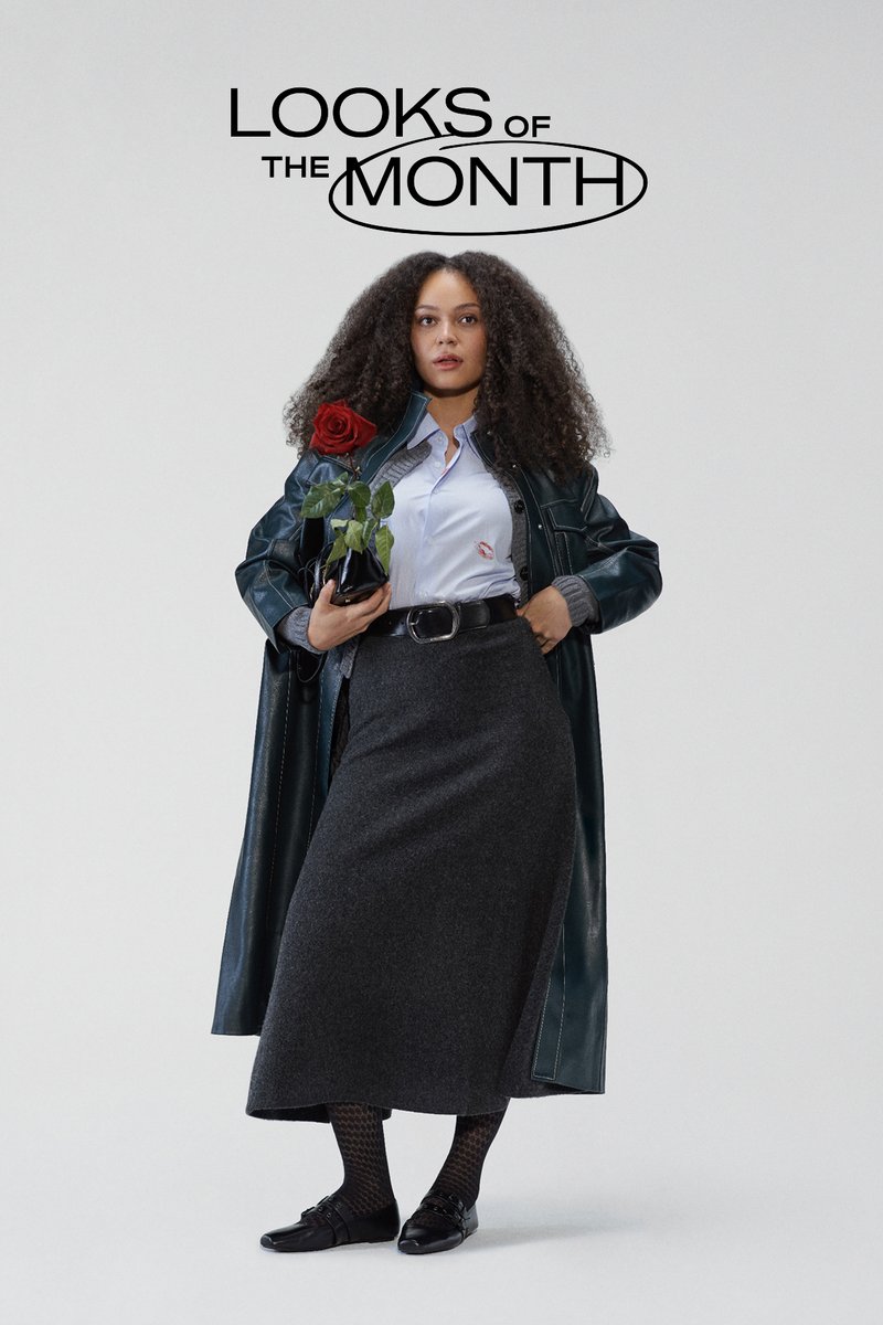 Model wearing a dark green leather coat, light blue shirt, grey skirt, and black shoes, holding a rose in a black pot.