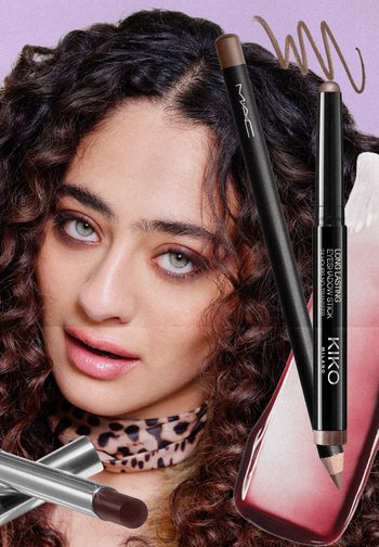 Femme aux cheveux bouclés portant un maquillage des yeux marron, entourée de bâtonnets et de traces d’ombres à paupières marron et mauve sur fond violet.