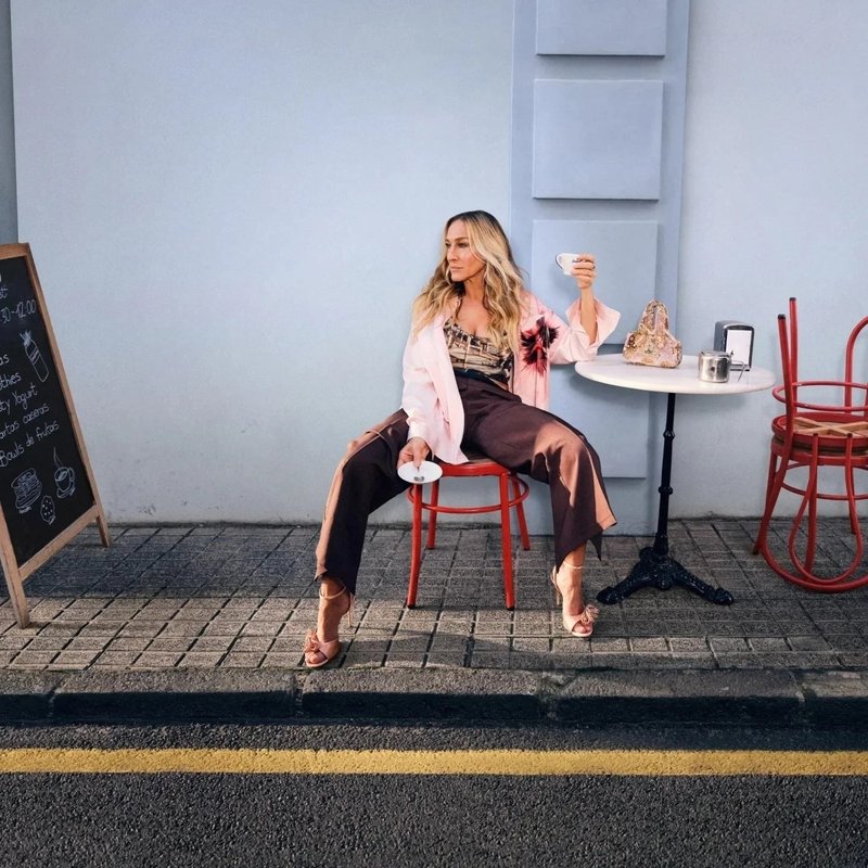 Roze jas, patroonbloes, bruine wijde broek, met een witte beker in de hand. Rode stoel en tafel tegen een lichtblauwe muur. Stoeptegels.