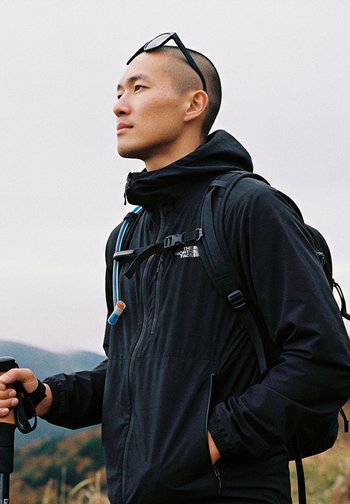 Giacca nera da esterno con colletto alto, dotata di zip frontale, tubo di idratazione attaccato e zaino. Tiene un bastone da trekking in mano.