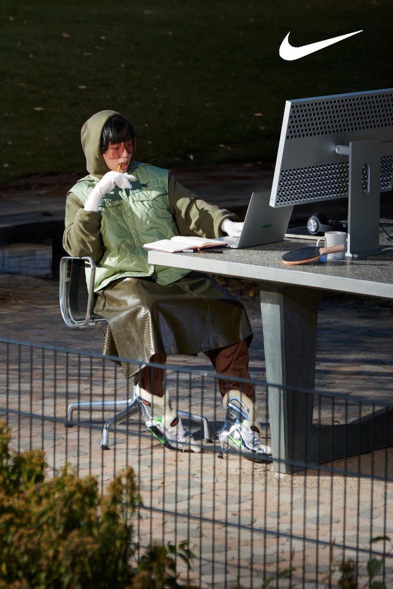 Een persoon in een groene vest en een bruine broek zit aan een tafel buiten, werkt op een laptop en houdt een potlood vast, met een notitieboekje in de buurt.