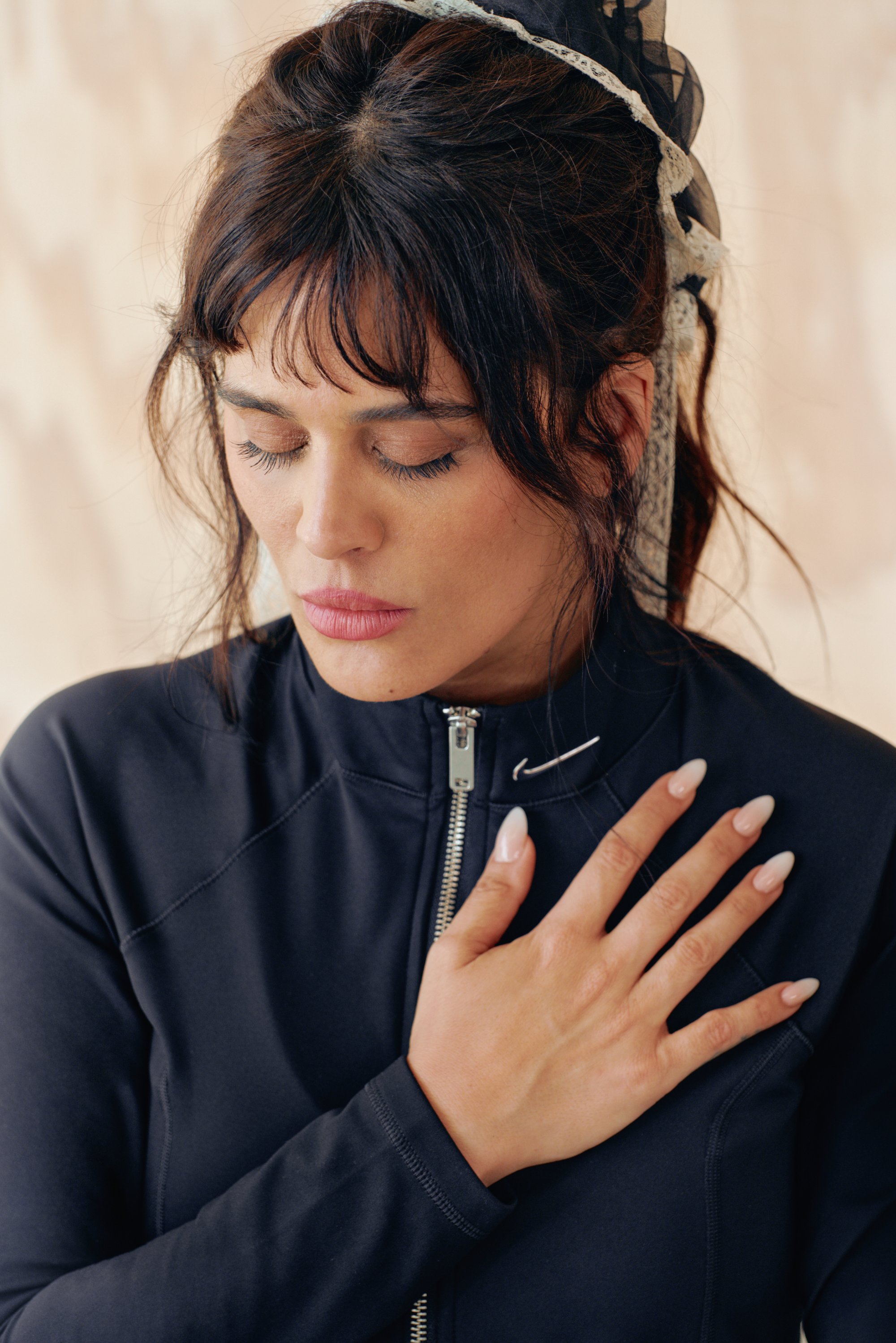 Chaqueta deportiva negra con cremallera y cuello alto, con un logo discreto. El modelo tiene el cabello oscuro despeinado y uñas de color claro, con la mano reposando sobre el pecho.