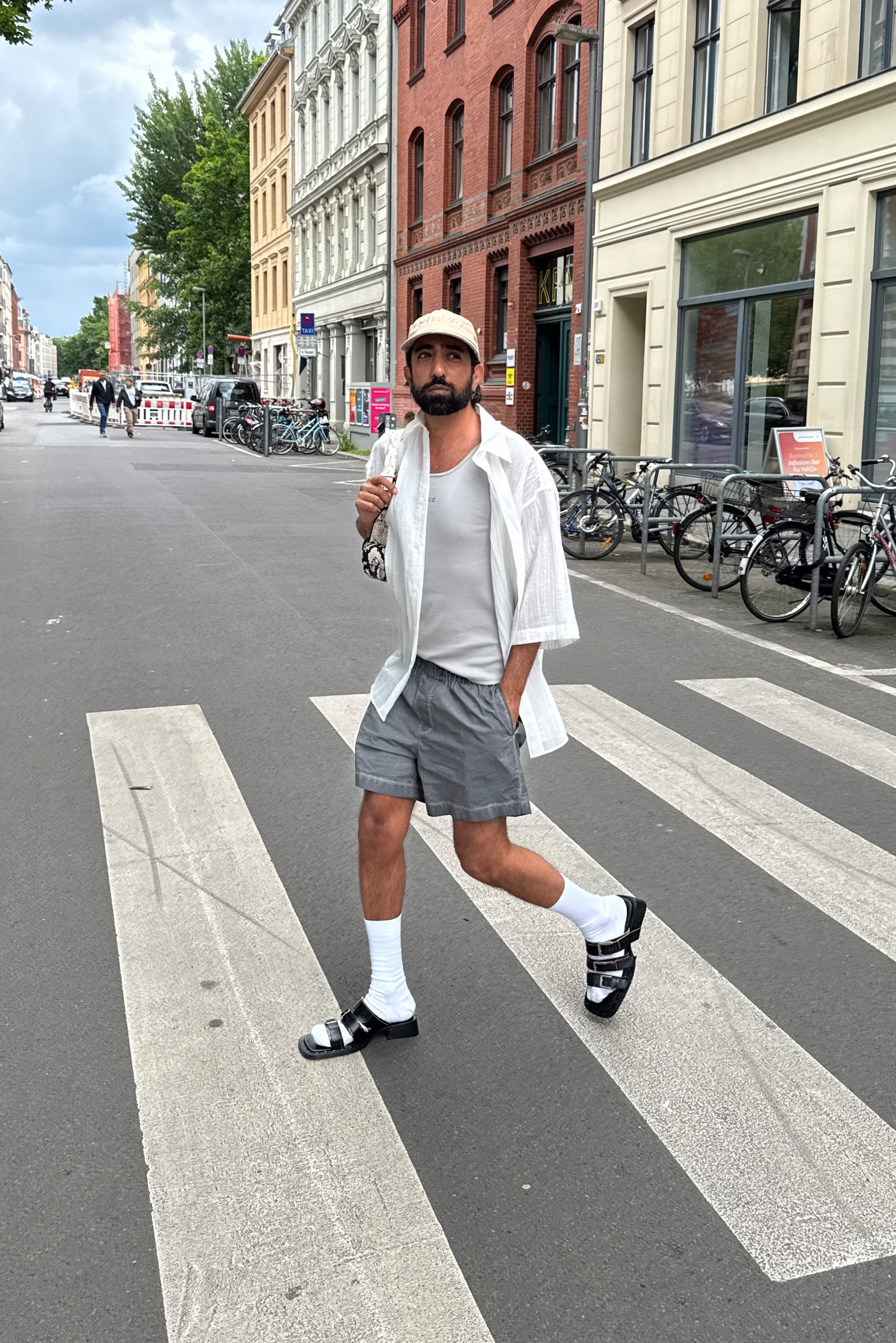 Un uomo cammina con sicurezza nel passaggio pedonale, indossando pantaloni corti, una canotta e una maglietta ampia alla moda, con un cappello e sandali. Le biciclette sono allineate lungo la strada.