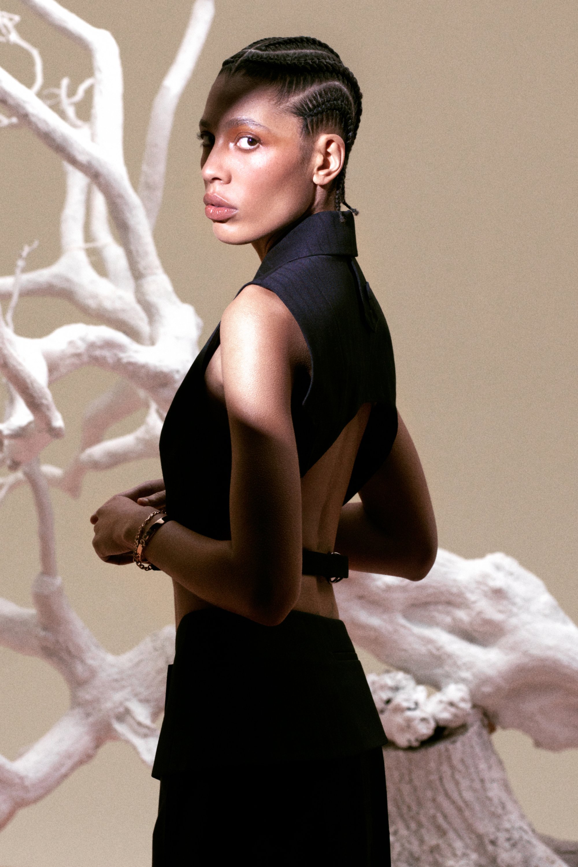 Modelo con cabello trenzado posa con un atuendo oscuro sin mangas, mostrando un corte en la espalda, contra un fondo claro con ramas abstractas blancas.