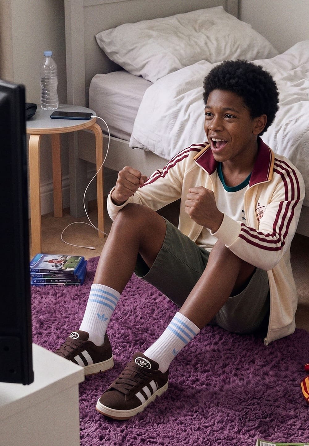 Boy in sportswear sitting on purple rug, excited and cheering while playing video games in a bedroom with a bed and side table nearby.