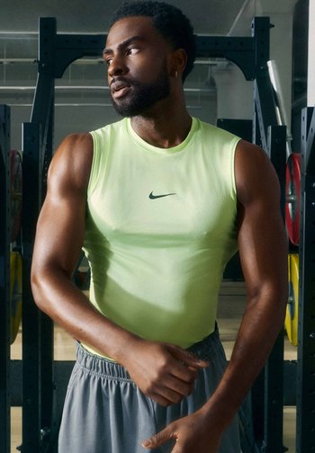 Muscular man in a bright sleeveless shirt adjusting his wristband in a gym with weightlifting equipment behind him.