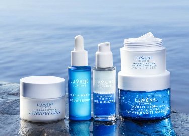 Cuatro productos de cuidado de la piel en envases azul y blanco de diferentes tamaños están dispuestos sobre una roca cerca del agua, mostrando un entorno natural y calmante.