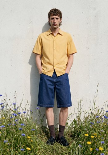 Man staat tegen een witte muur, draagt een geel shirt met korte mouwen, knielange blauwe spijkerbroekshorts, bruine sokken en zwarte schoenen, met wilde bloemen in de buurt.