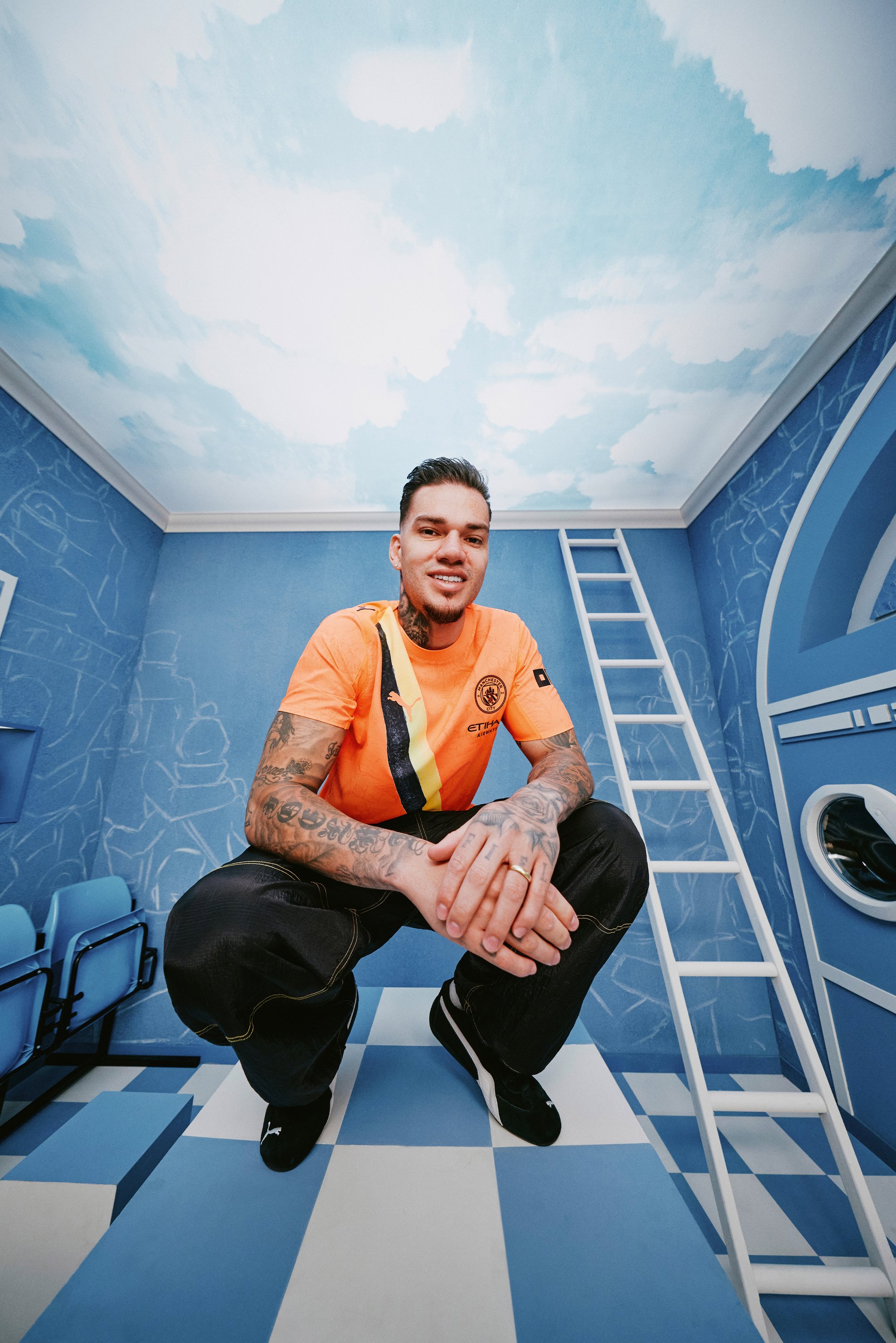 A man in an orange shirt with black and yellow accents poses in a blue room with clouds on the ceiling and a ladder in the background.