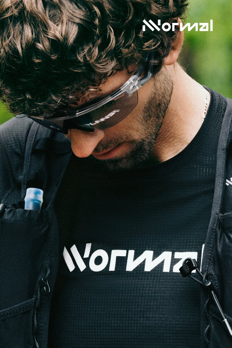A man with curly hair, wearing sunglasses and a black shirt, gazes down, showcasing a logo and a water bottle in a backpack pocket.
