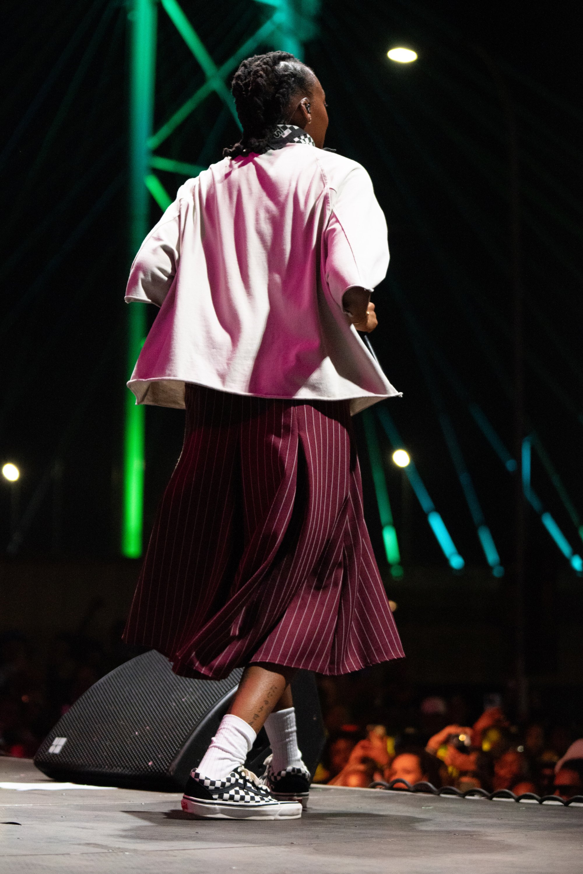 A performer in a loose, patterned outfit dances on stage, surrounded by a vibrant audience and illuminated by colorful stage lights.