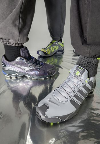 Dois pares de ténis desportivos: um Nike cinzento com couro texturizado, detalhes verdes e um logótipo proeminente; o outro Mizuno metálico roxo com detalhes em rede.