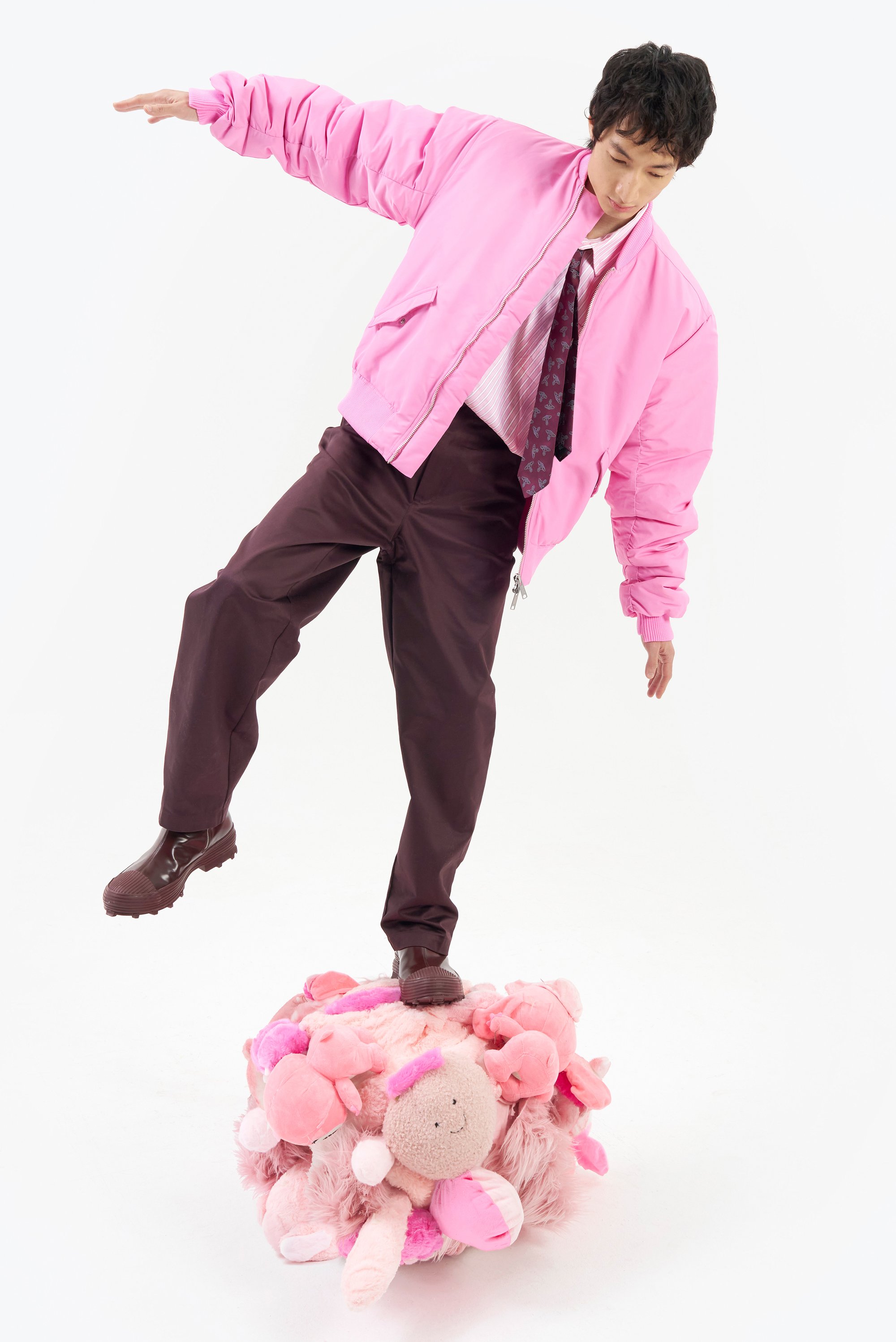 Una persona se equilibra sobre un montículo esponjoso y rosa adornado con juguetes de peluche, vistiendo una chaqueta bomber rosa, una camiseta blanca y pantalones marrones.