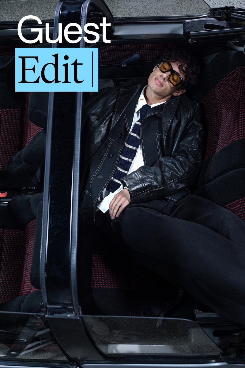 Una persona se relaja en un coche, vistiendo una chaqueta de cuero y gafas de sol, con una corbata a rayas con estilo. El interior del coche cuenta con asientos con acentos rojos.