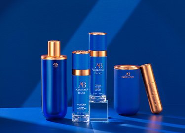 Cztery produkty do pielęgnacji skóry w niebieskich butelkach ze złotymi akcentami, w tym spray, dwa sera i tuba, ułożone na niebieskiej powierzchni.