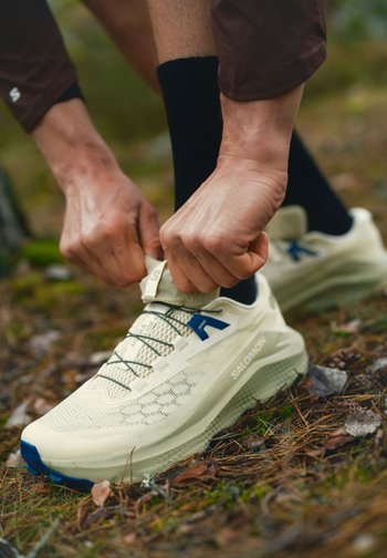 Hender som knyter lisser på en beige Salomon terrengløpesko, brukt med svarte sokker, utendørs på en skogbunn dekket med blader og gress.