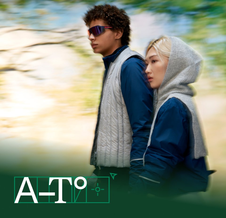 Two young adults walk side by side against a blurred natural background. The man wears sunglasses and a light vest, while the woman has a hoodie.