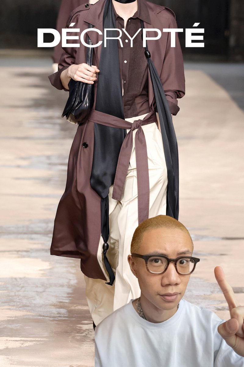 Personne aux cheveux blonds courts et portant des lunettes lève l'index devant un mannequin sur le podium, vêtu d'un manteau marron, d'une écharpe noire et d'un pantalon crème.