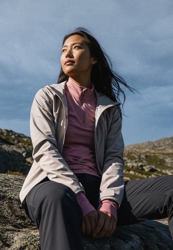 Giacca grigia chiara con zip sopra una maglietta rosa a maniche lunghe, abbinata a pantaloni scuri. Tessuto testurizzato e vestibilità casual, ambientazione all'aperto.
