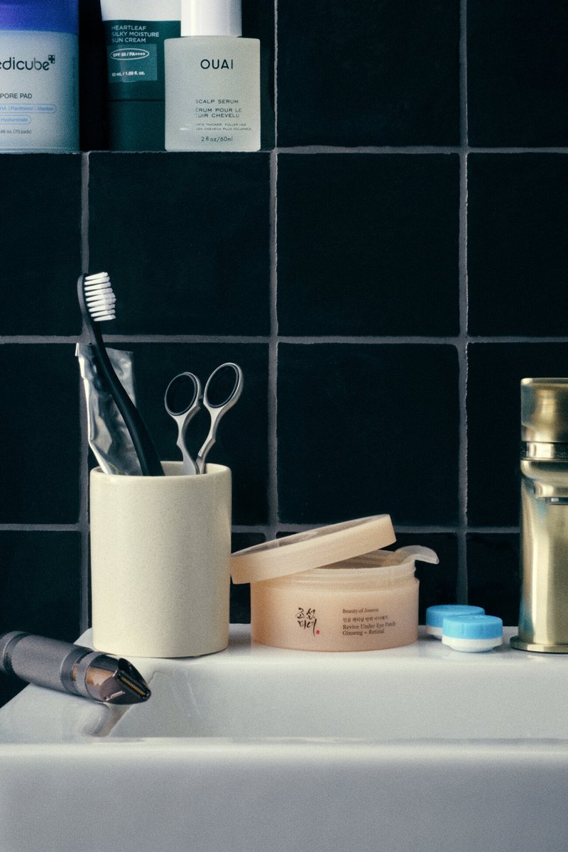 Brosse à dents, ciseaux et dentifrice dans une tasse, pot de soins de la peau ouvert, étui de lentilles de contact et tondeuse sur un lavabo blanc avec un mur carrelé noir.