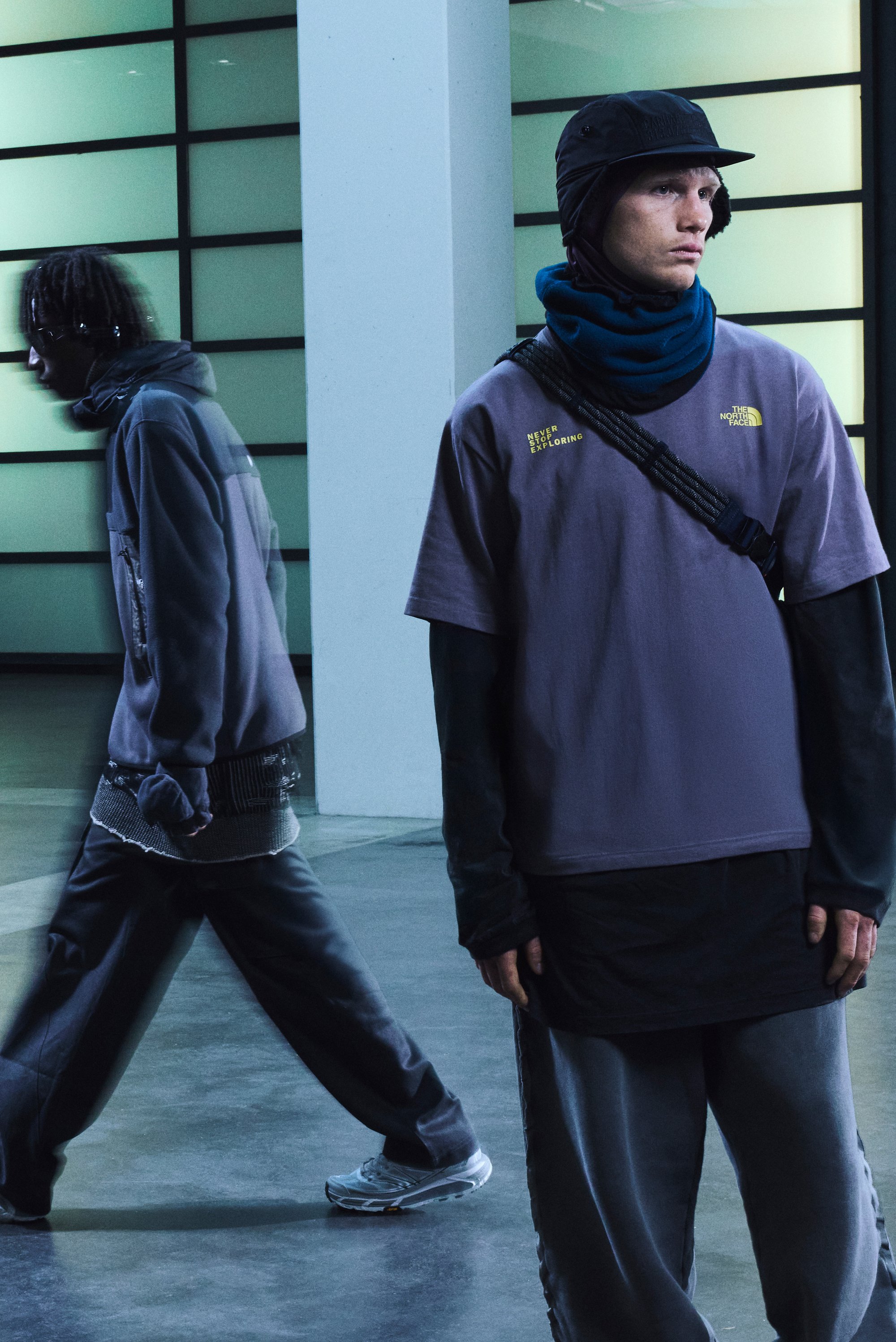 Due modelli indossano abbigliamento urbano. Uno ha una giacca scura e un cappellino nero; l'altro indossa una maglietta viola e pantaloni scuri. Entrambi presentano design stratificati.