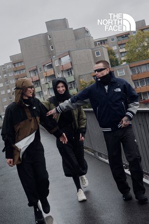 Trois personnes marchent le long d'une rue urbaine humide. Elles portent des vestes résistantes aux intempéries, des pantalons noirs et des chaussures de sport. Les couleurs dominantes sont le vert olive, le noir et le beige.