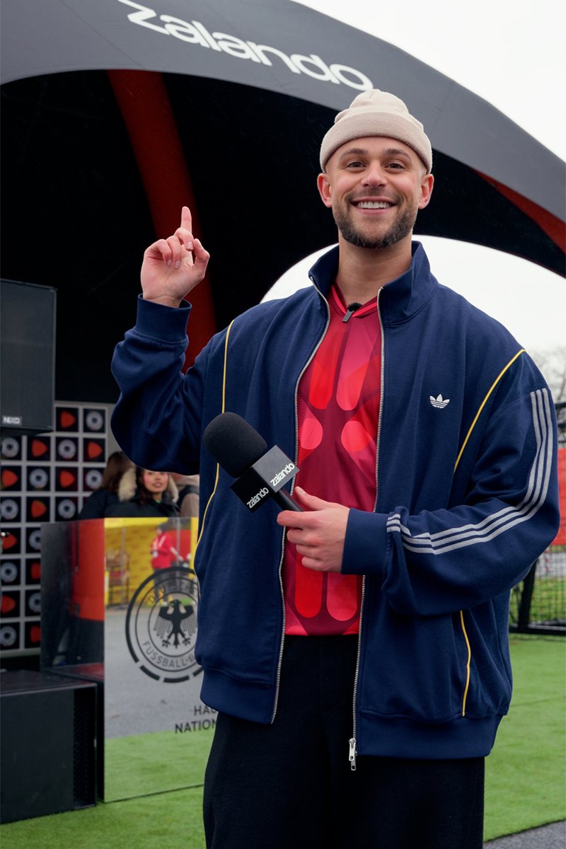 Smiling man in beanie and sportswear holds Zalando microphone, points up under Zalando-branded arch at outdoor event.