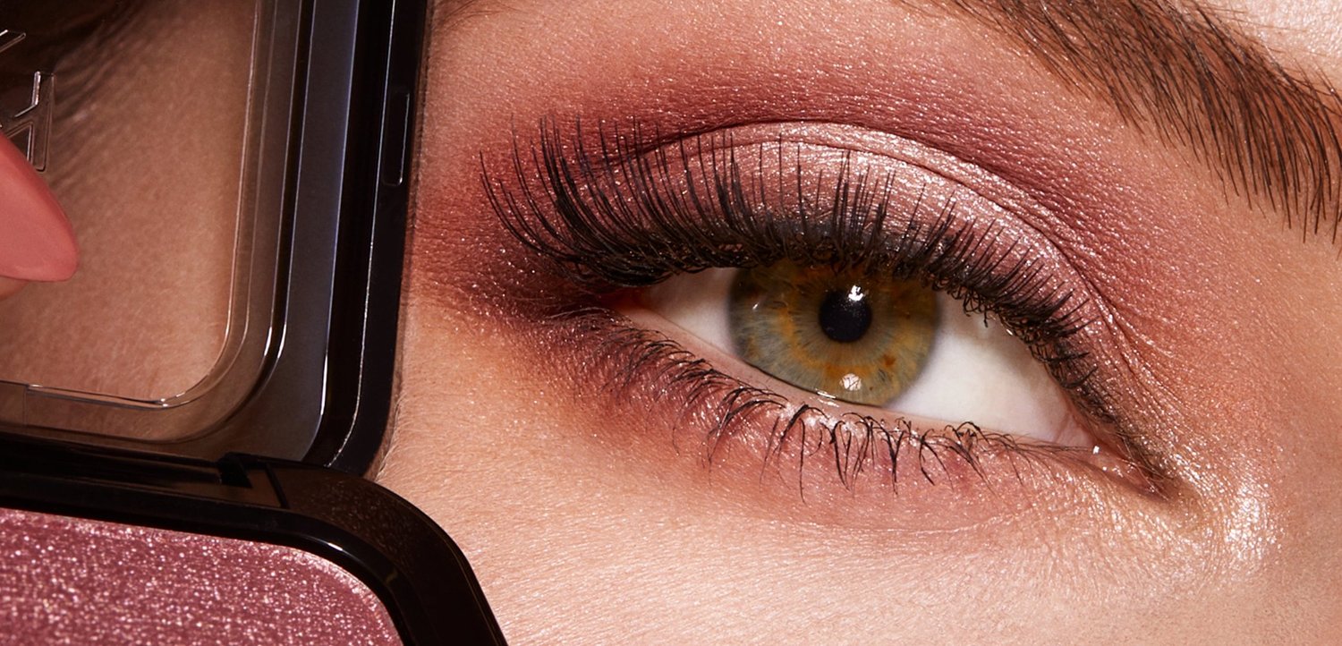 Nahaufnahme eines haselnussbraunen Auges mit rosa Lidschatten, dichten Wimpern und einem Make-up-Compact, der in der Nähe schimmernden rosa Lidschatten aufträgt.