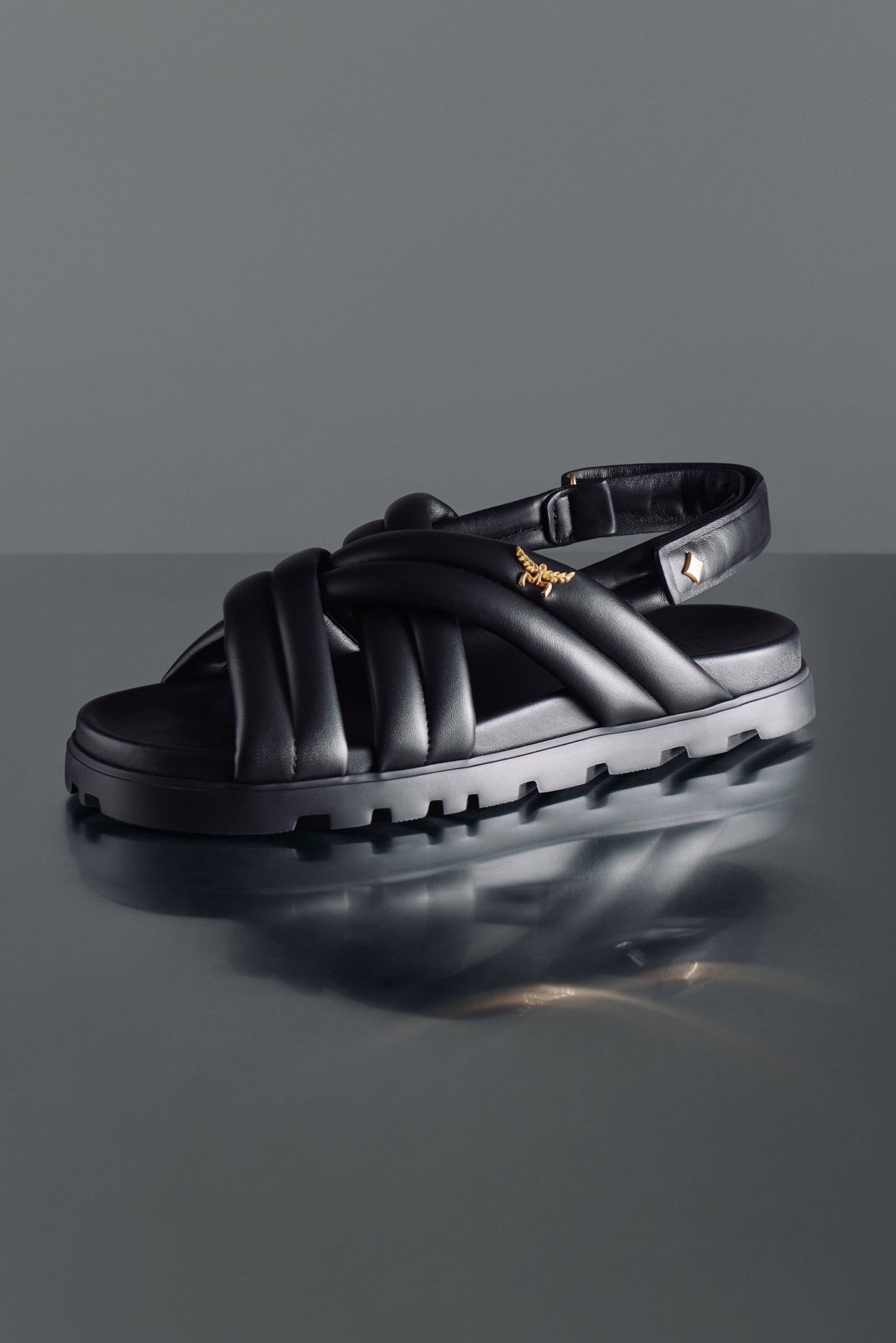 Sandaleta negra con gruesas correas trenzadas y trasera ajustable, que presenta un sutil logo dorado. Superficie reflectante en la parte inferior.