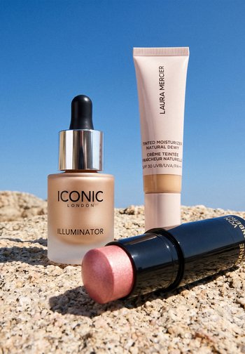 Flacon compte-gouttes iconique London Illuminator, tube de crème hydratante teintée Laura Mercier et enlumineur stick rose L'Oréal sur une surface en pierre sous un ciel bleu.