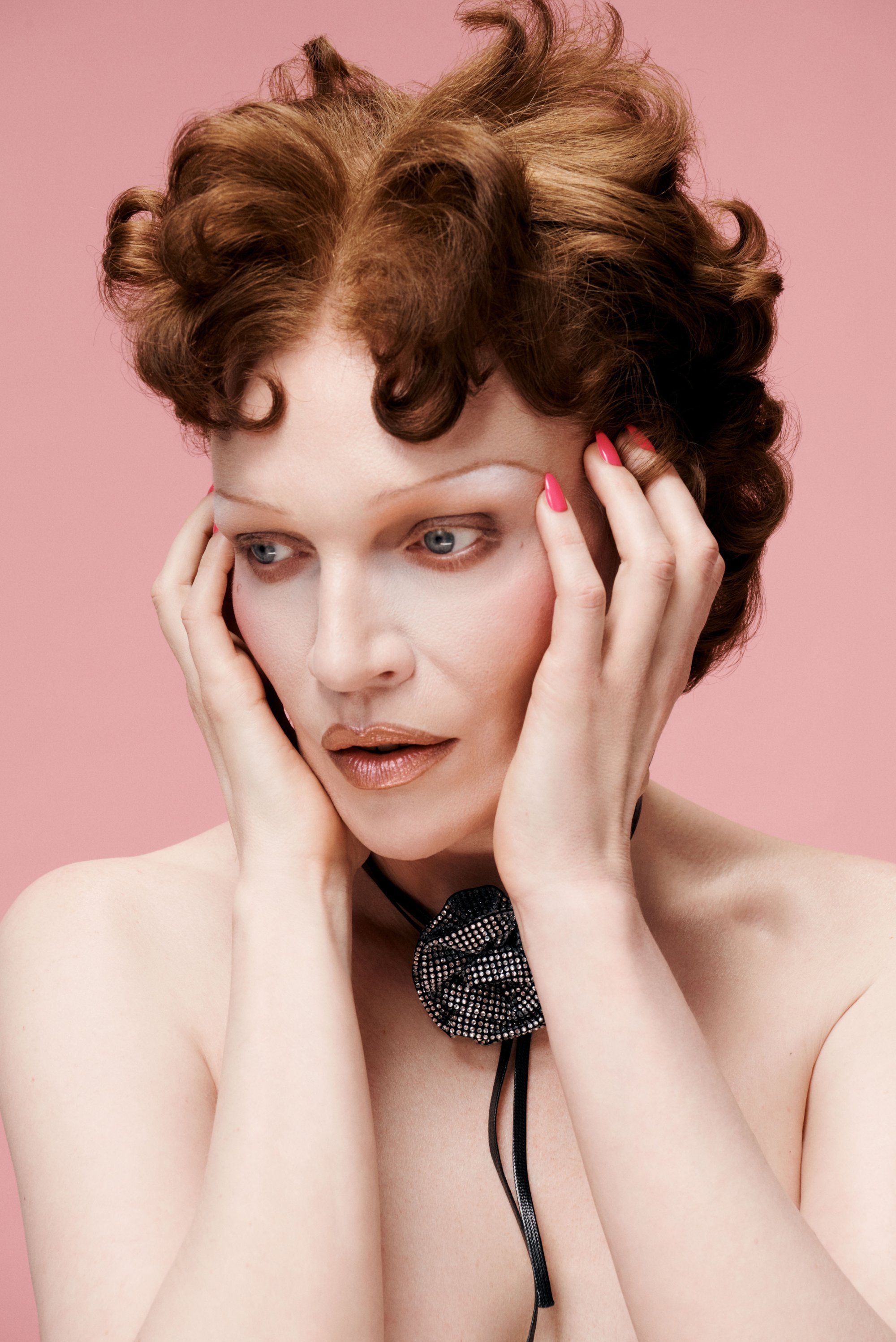 Primer plano de una persona con cabello rizado y rojo, que lleva un choker de encaje negro con un diseño floral. La piel parece clara, con un maquillaje suave y un fondo rosa.