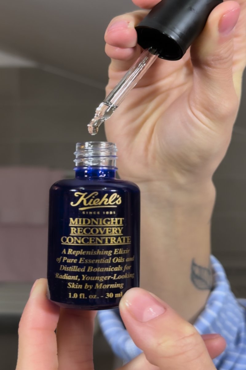 Hand holding a Kiehl's Midnight Recovery Concentrate bottle with a dropper releasing clear liquid above the open bottle.