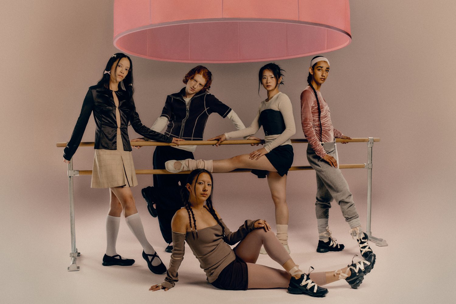 Vijf dansers poseren rond een balletbar, terwijl ze diverse outfits en stijlen tonen. Boven hen hangt een grote roze lampenkap in een minimalistische setting.